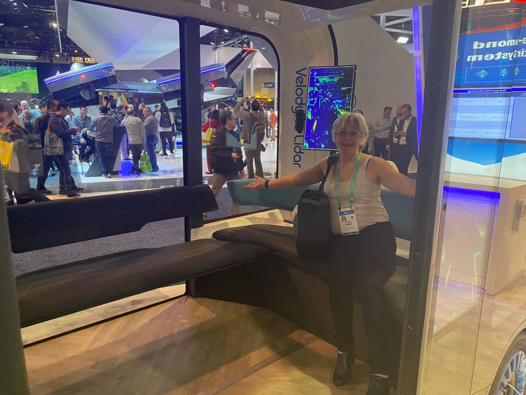 A person sitting in a futuristic vehicle display at the Consumer Electronics Show, surrounded by attendees and technology exhibits in the background.