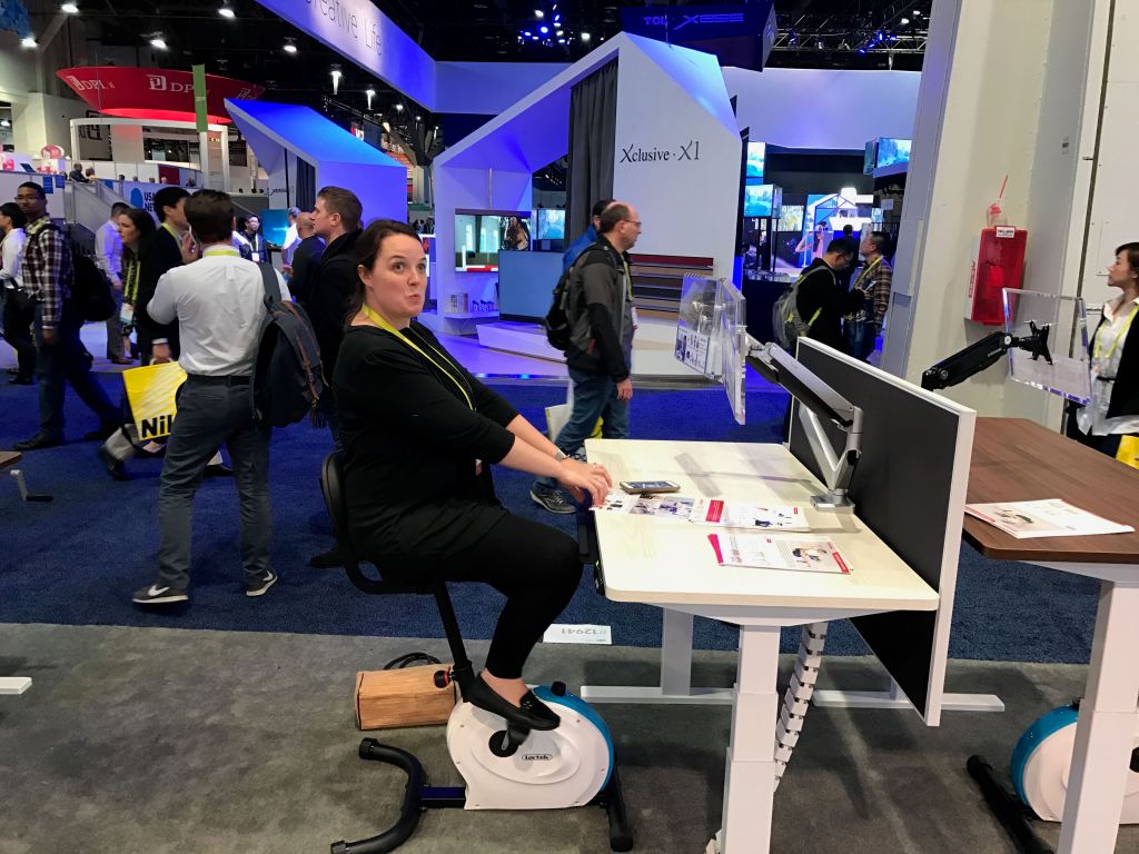 A woman sitting on an exercise bike at a technology exhibition, interacting with a desk setup featuring computer monitors as people walk by in a busy convention hall.