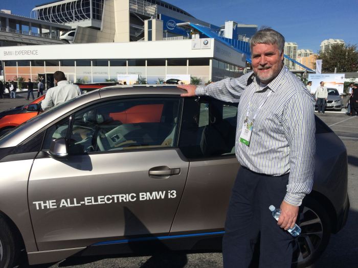 A man standing next to an all-electric BMW i3 at the Consumer Electronics Show, with the BMW Experience pavilion in the background.