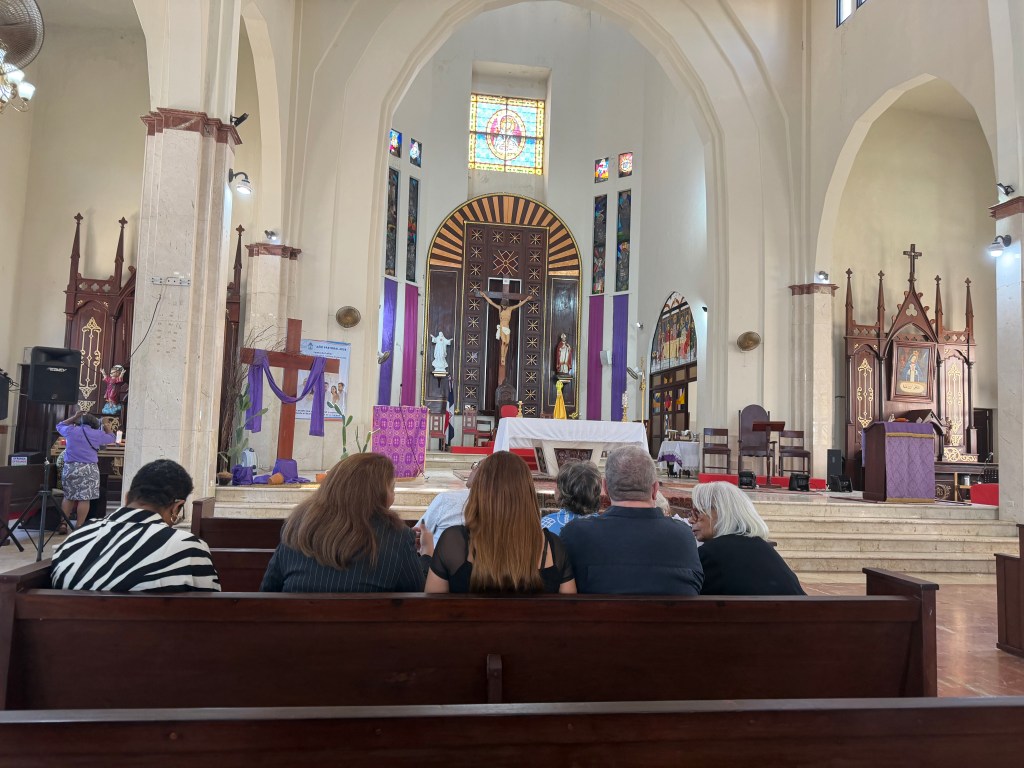 Interior view of a church with worshippers seated on wooden benches, facing the altar adorned with religious decorations and colorful stained glass windows.