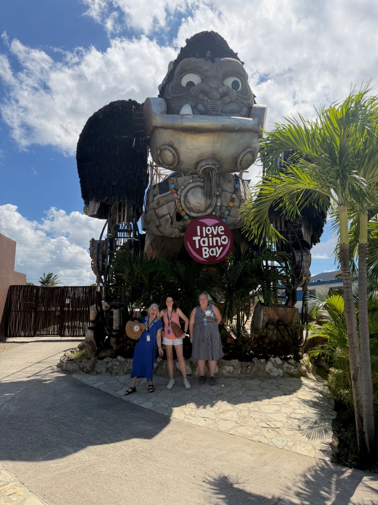 Three women posing in front of a large colorful statue with the text 'I love Taino Bay' surrounded by palm plants and a blue sky.