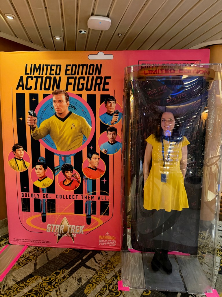 A display featuring a limited edition Star Trek action figure box with illustrations of characters, alongside a person in a yellow dress standing behind a transparent cover, mimicking the action figure presentation.