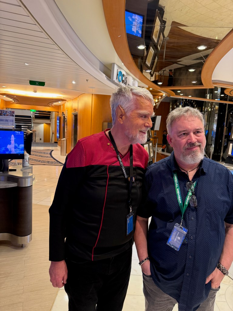 Two men standing together in a cruise ship interior, one wearing a Starfleet uniform top and the other in a dark blue shirt with glasses. They appear to be engaged in conversation.