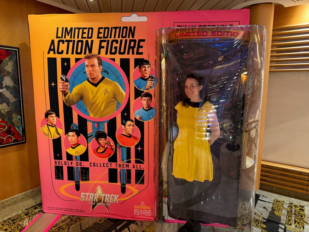 A woman stands in a plastic display resembling an action figure packaging, dressed in a yellow dress, next to a colorful poster featuring characters from Star Trek, promoting a limited edition action figure.