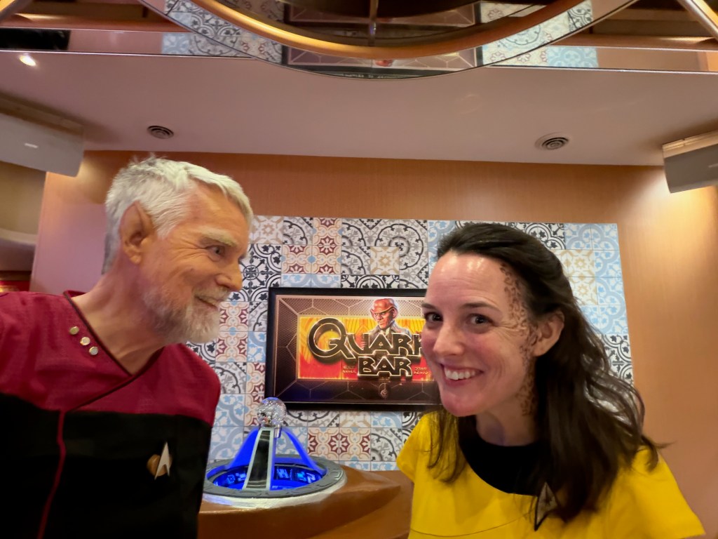 Two people dressed in Star Trek-themed costumes smiling at each other in a bar setting, with a colorful backdrop featuring a sign for 'Quark's Bar'.