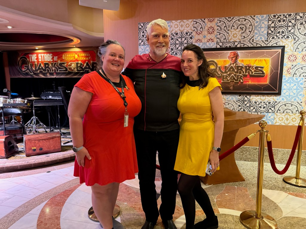 Three friends posing together in front of a colorful backdrop featuring 'Quark's Bar'. One is wearing a red dress, another is in a black Star Trek uniform, and the last is dressed in a yellow dress.