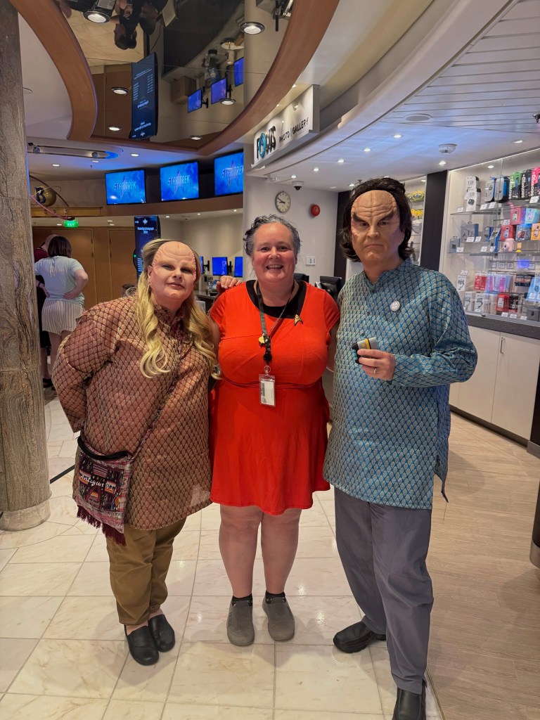 Two individuals in costume, resembling extraterrestrial characters, stand beside a woman wearing a red dress in a cruise ship environment with electronic displays in the background.
