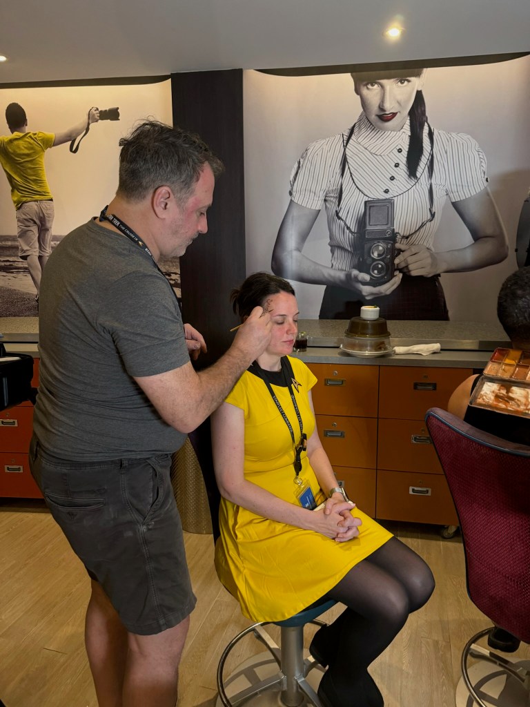 A makeup artist applying makeup to a woman sitting on a stool in a professional setting. The woman is wearing a yellow dress, and there's a backdrop featuring a vintage photography theme.