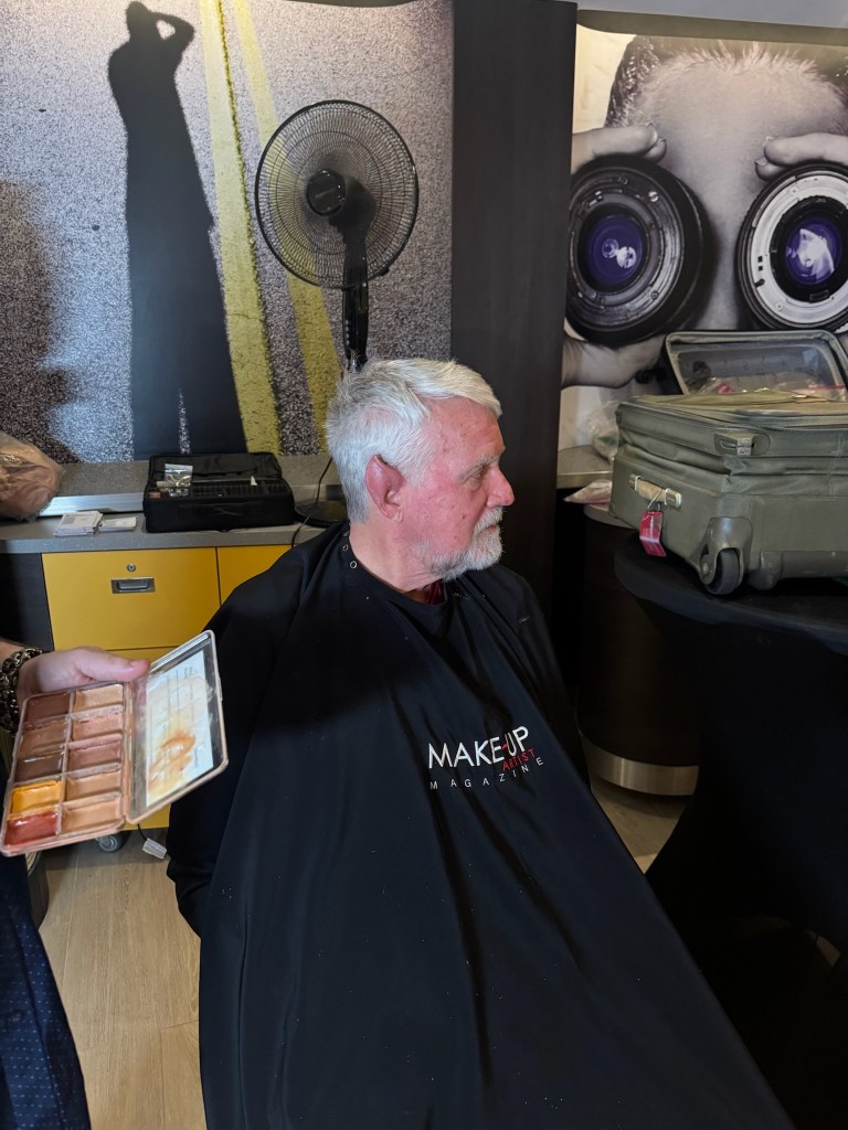 A man with gray hair sits in a makeup chair wearing a black cape with 'MAKEUP MAGAZINE' printed on it. A hand holds a palette of makeup colors near him, and a fan is visible in the background.