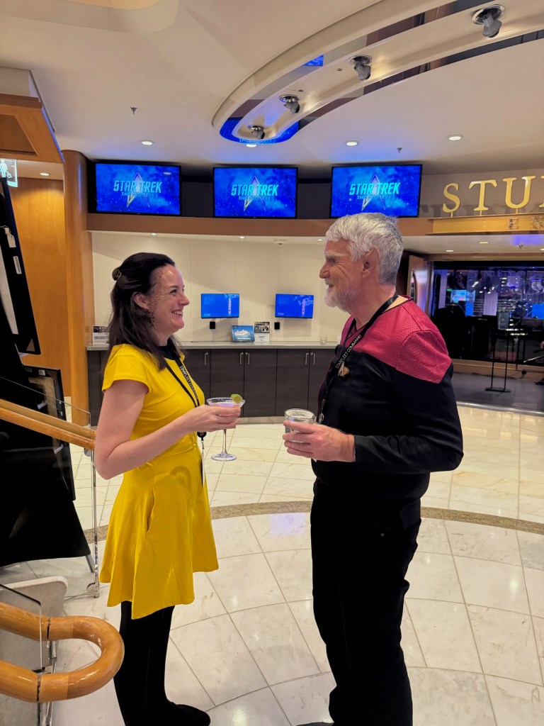 A woman in a yellow dress and a man in a black outfit with a red collar are smiling and chatting while holding cocktails in a stylish indoor setting with Star Trek-themed decor in the background.